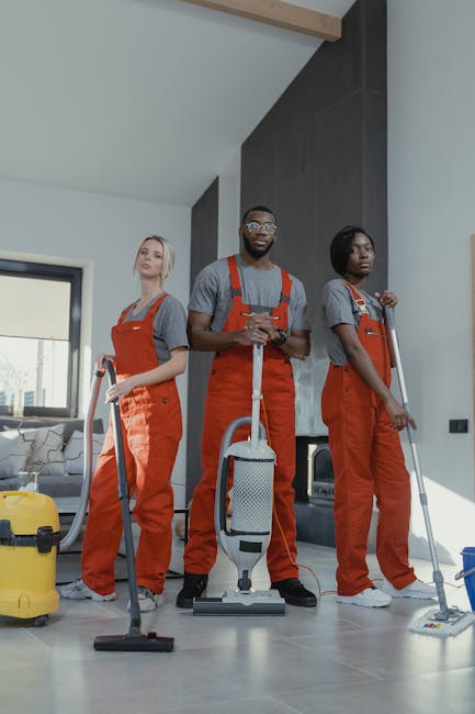 Two professional cleaners in orange uniforms are depicted in a modern living room with white walls and large windows. The male cleaner is holding a cleaning tool and a cloth, preparing for surface cle